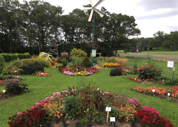 Small formal garden beds full of flowers, divided by neat lawn. A small windmill and tall trees stand in the background.