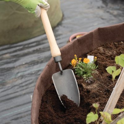 hands wearing green garden gloves gripping a trowel and digging in a large planter