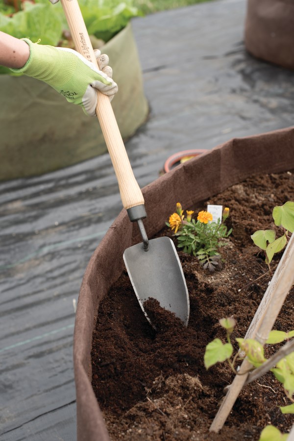 hands wearing green garden gloves gripping a trowel and digging in a large planter