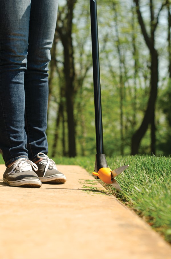 close-up image of a person's lower half wearing jeans and sneakers and using long-handled shears to cut grass along a walkway