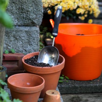 Terracotta pots, one filled with soil and a hand trowel, and an orange bucket full of soil in front of a pot of yellow mums