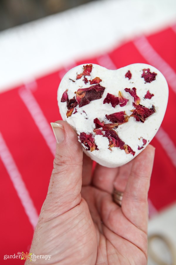 Woman holding a heart-shaped bath bomb with dried rose petals inside.