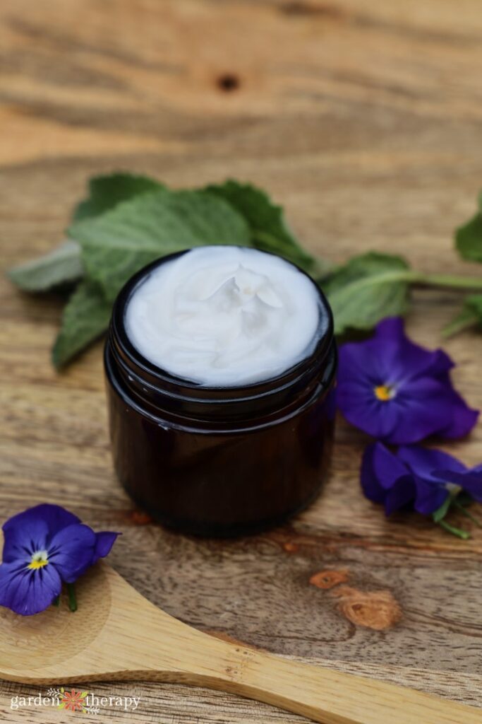 Whipped hand cream in an amber jar surrounded by purple flowers.