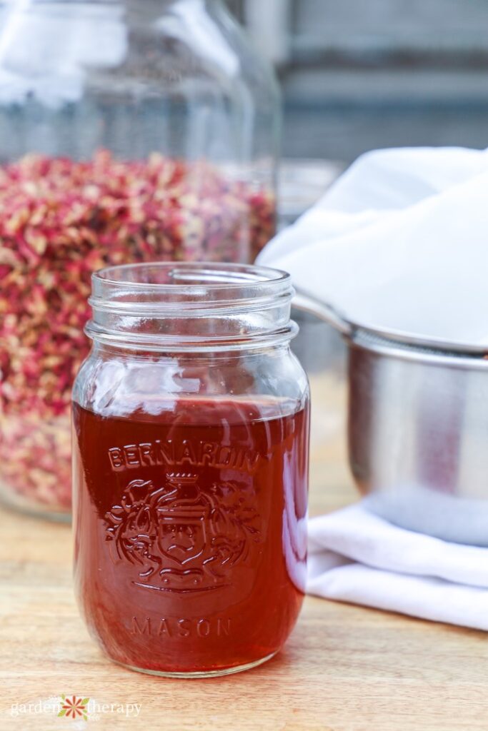 rose water in a mason jar in front of a pot and a jar of dried rose petals