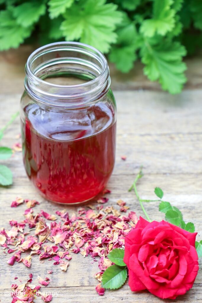 Mason jar filled with pink rose water, sitting on top of wooden table with dried rose petals and a fresh rose